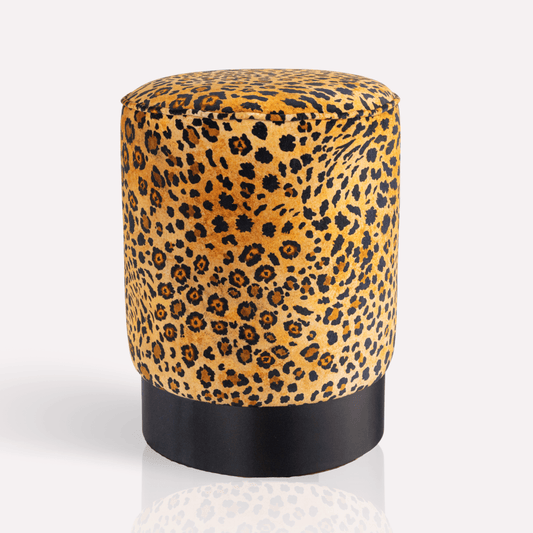 A cylindrical velvet pouffe featuring an opulent gold and black leopard-spot pattern, elegantly perched on a sleek black brass base.