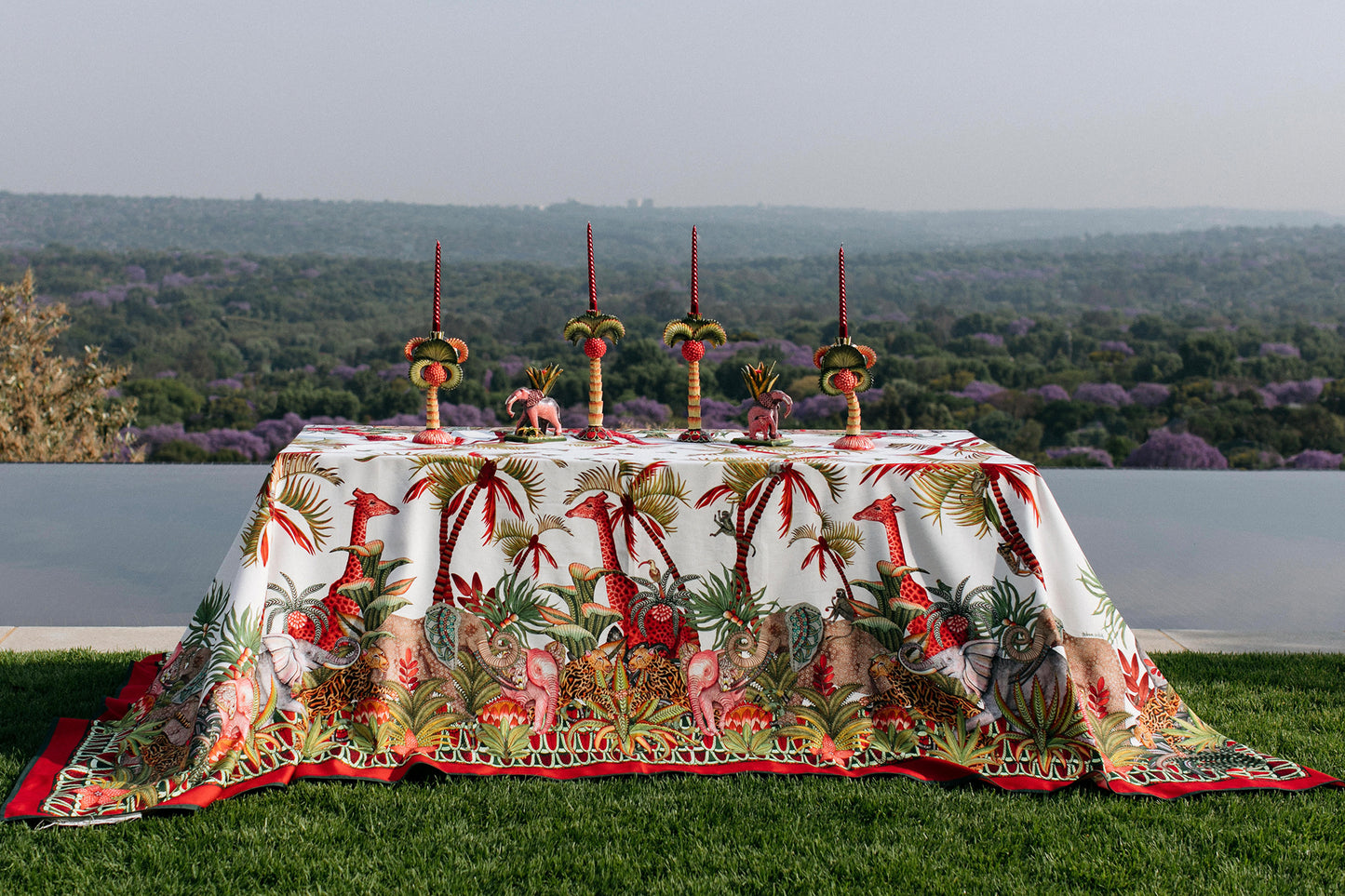 A scenic display of the Palm Parade tablecloth draped over a table set against a breathtaking outdoor backdrop, showcasing its lively colours and festive design.