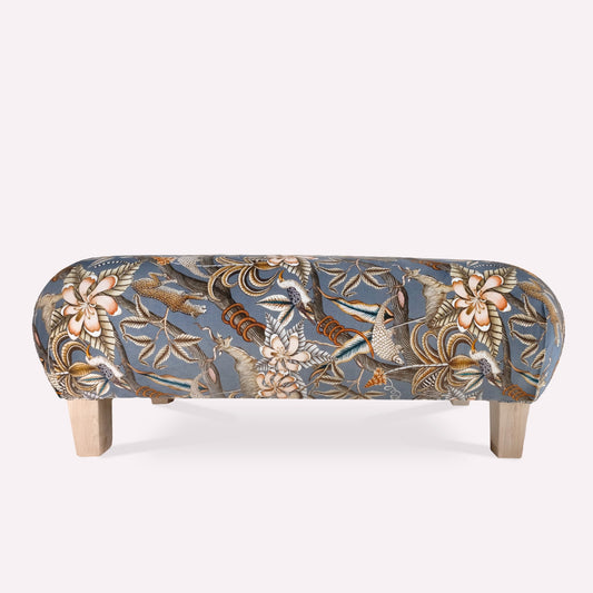 A luxurious rectangular ottoman upholstered in Pangolin Park Ash velvet, featuring an intricate golden botanical design against a deep blue background.