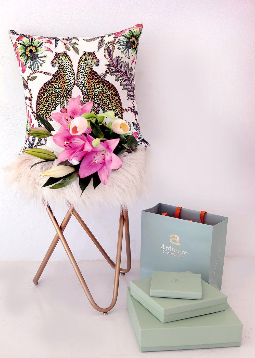 Styled view of the Lovebird Leopards Parakeet cushion cover on a chair with floral decor, paired with a gift box.