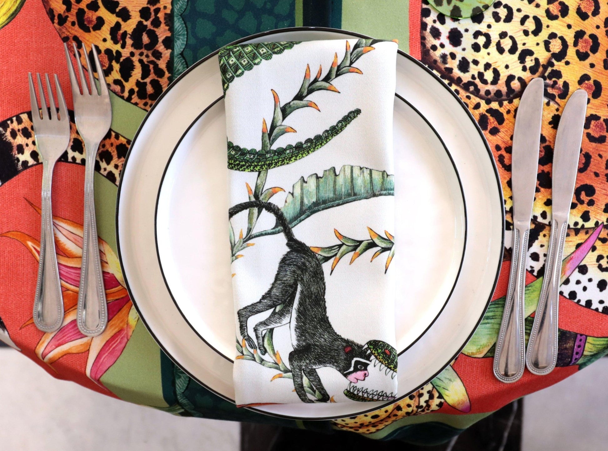 A neatly arranged stack of elegant plates on top of the Monkey Paradise Square Tablecloth, highlighting the contrast between the tableware and the vibrant fabric design.