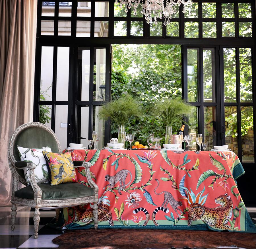The Monkey Paradise Tablecloth in Coral used in a stylish indoor setup, draped over a seating area with plush cushions.