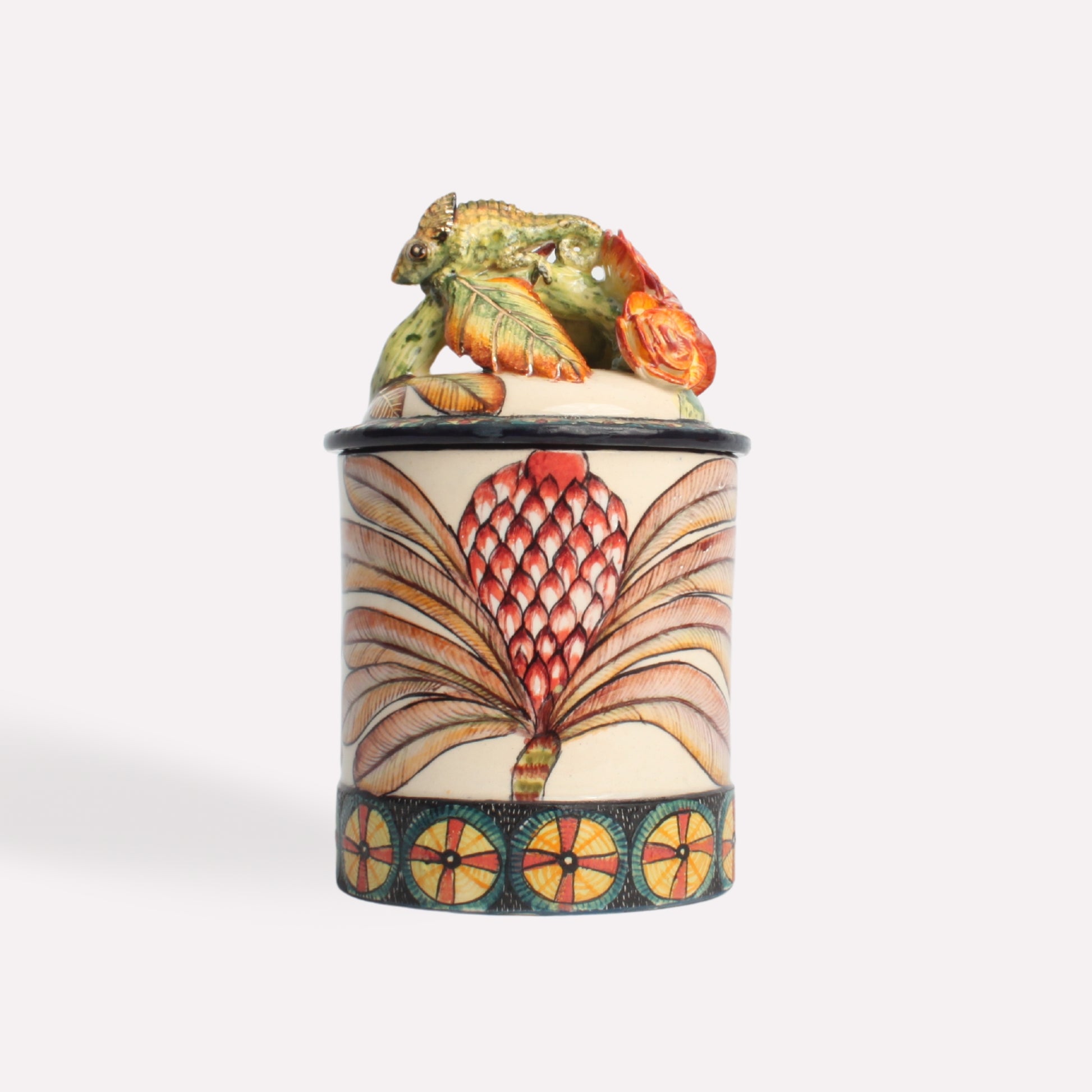 A close-up of the ceramic candle jar, highlighting the detailed chameleon figurine resting on the lid. The body of the jar is adorned with elegant geometric patterns and earthy colours.
