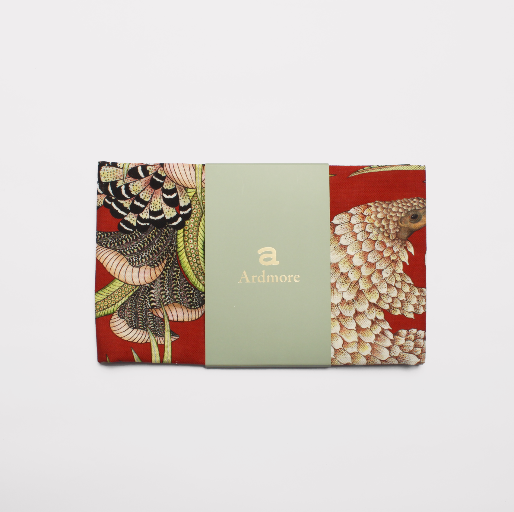 A packaged tea towel with a visible preview of the pangolin and botanical artwork, set against a striking red fabric background.