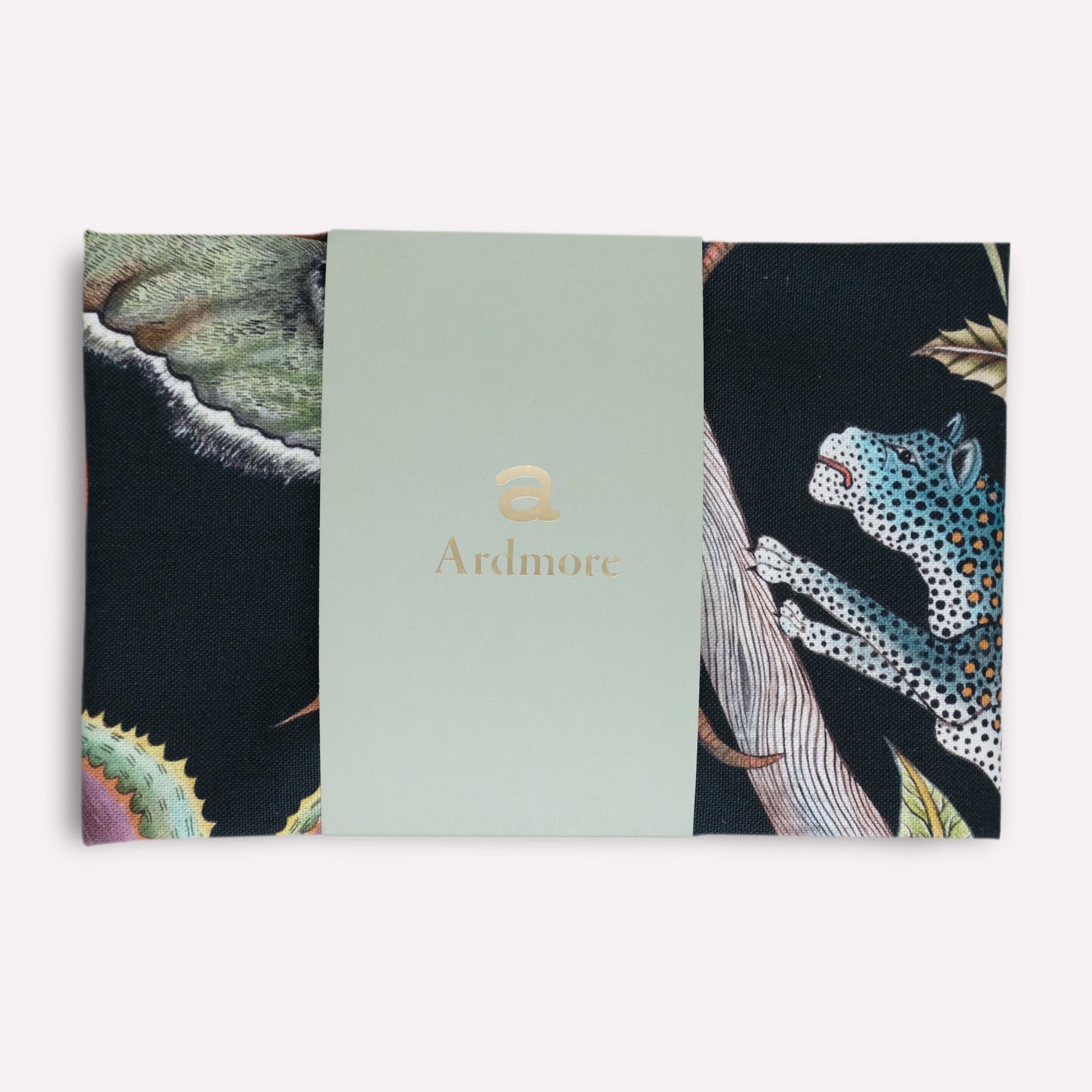 A tea towel in its packaging, displaying a sophisticated safari-themed artwork with lush foliage and striking animal illustrations.