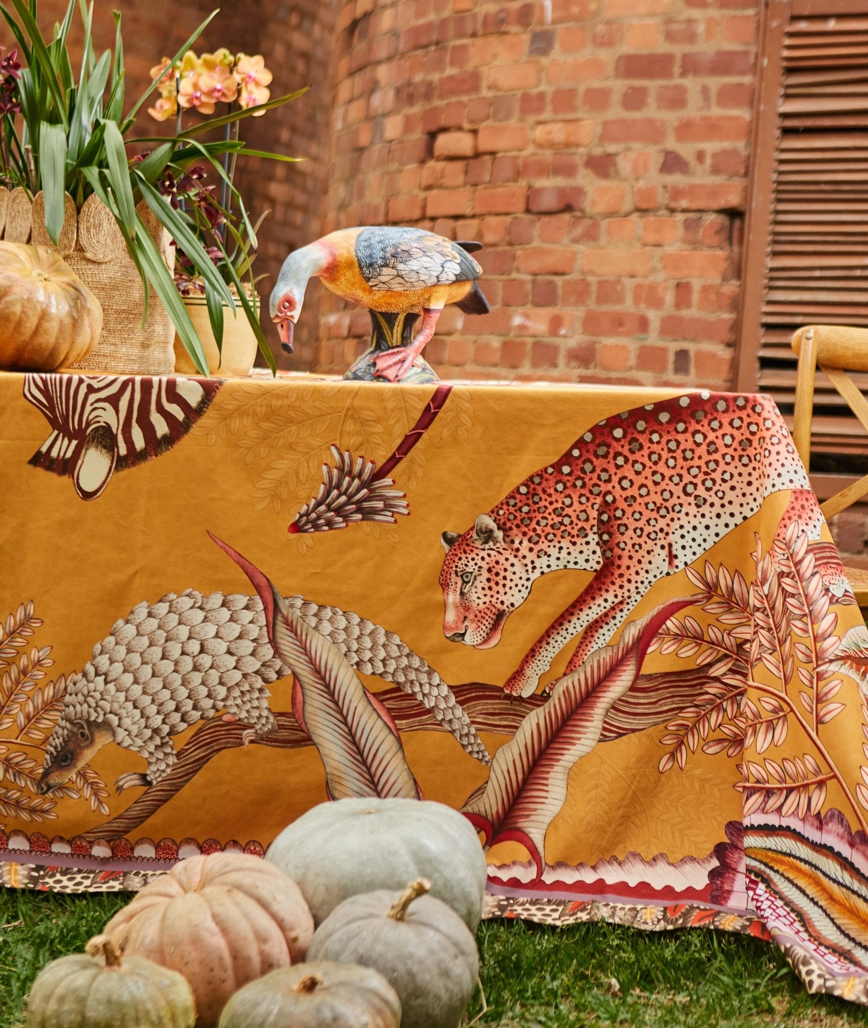 A dining table set outdoors, draped with the Pangolin Park tablecloth, surrounded by elegant tableware and floral décor.